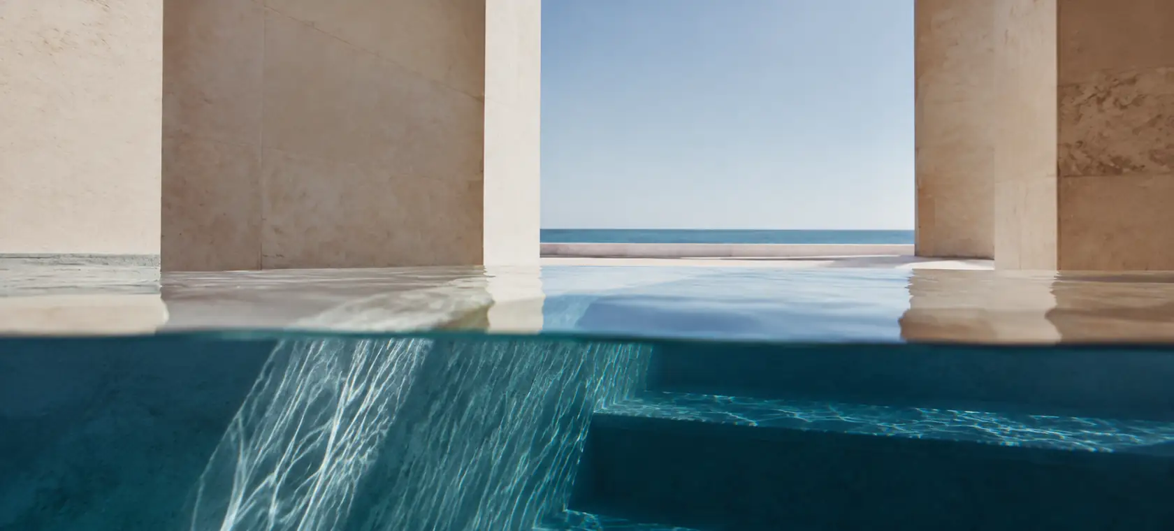 Vista al nivel del agua que divide un patio de piedra claro y la geometría azul sumergida de una piscina
